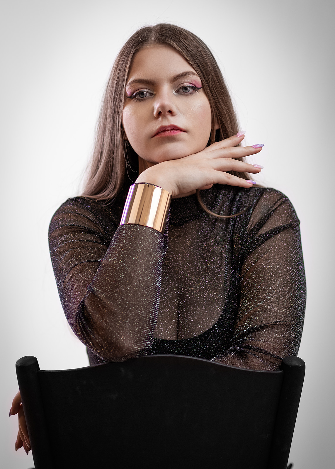 Young woman wearing a black sheer top and gold bracelet posing against a light background.