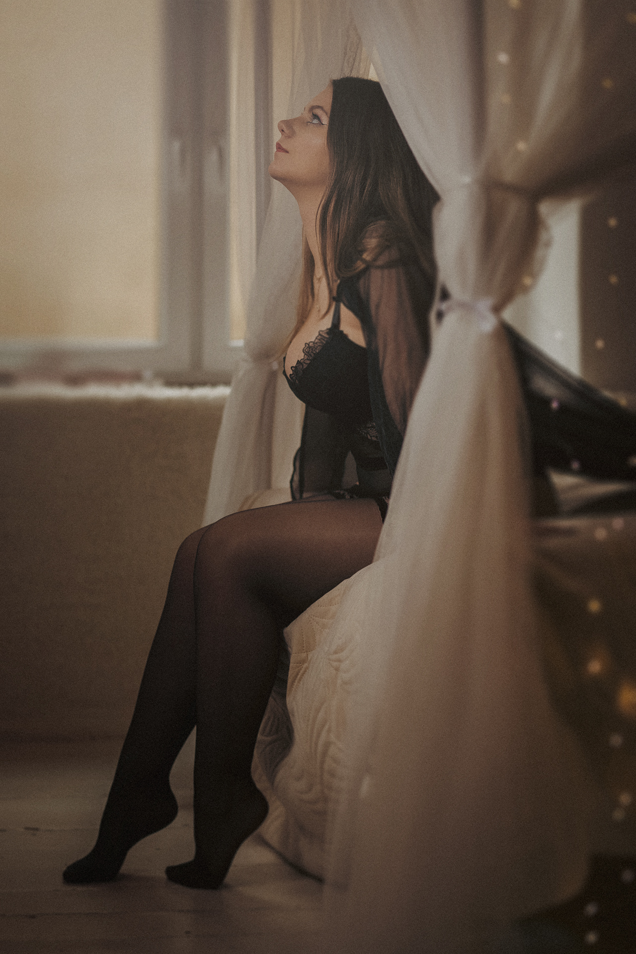 Woman sitting beside sheer curtains wearing elegant black lingerie.