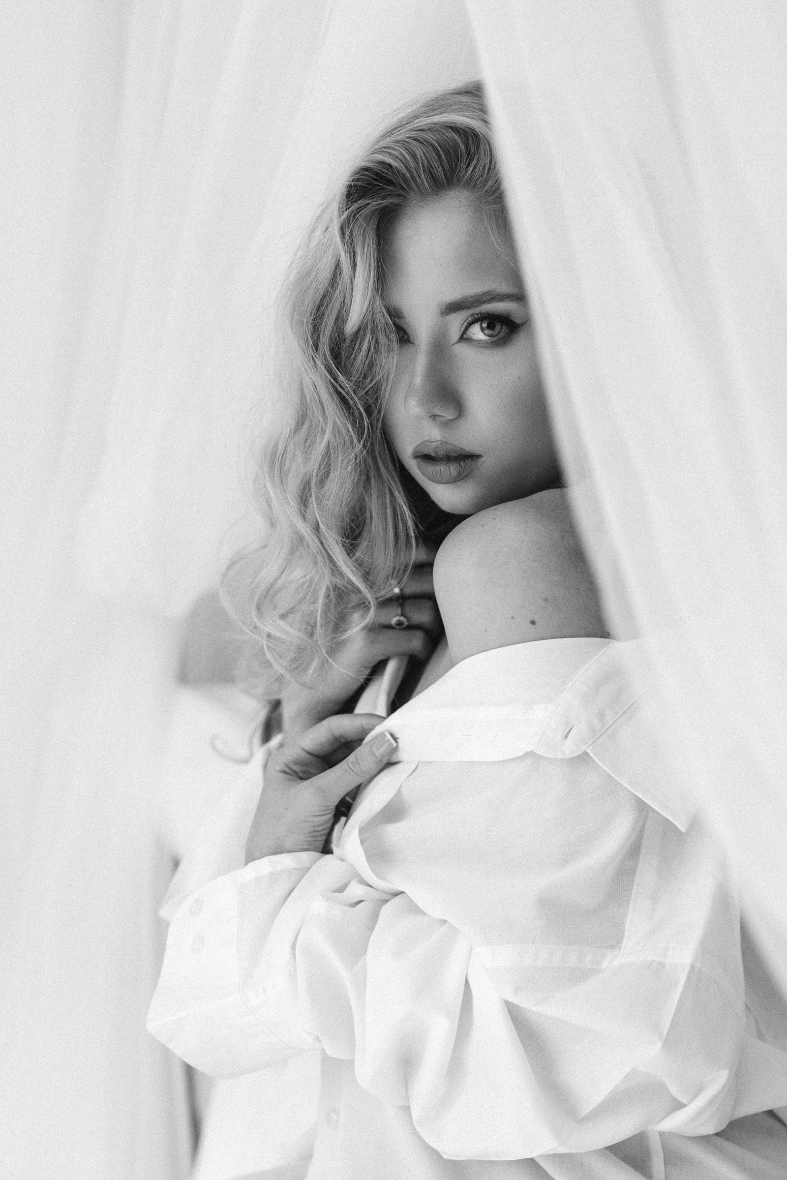 Black and white portrait of a blonde woman looking through sheer white curtains.