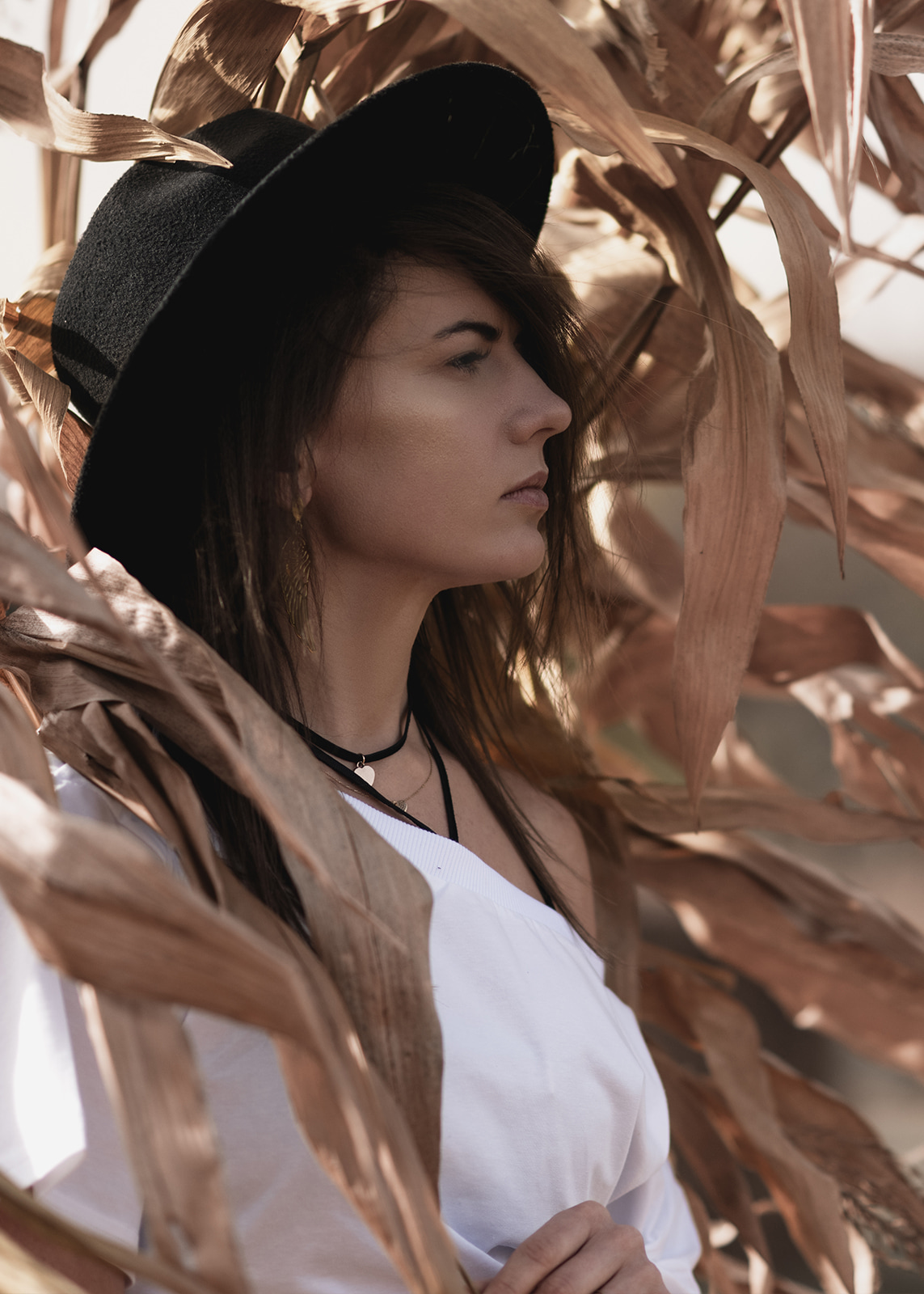 Side profile portrait of a woman wearing a black hat among dried corn leaves.