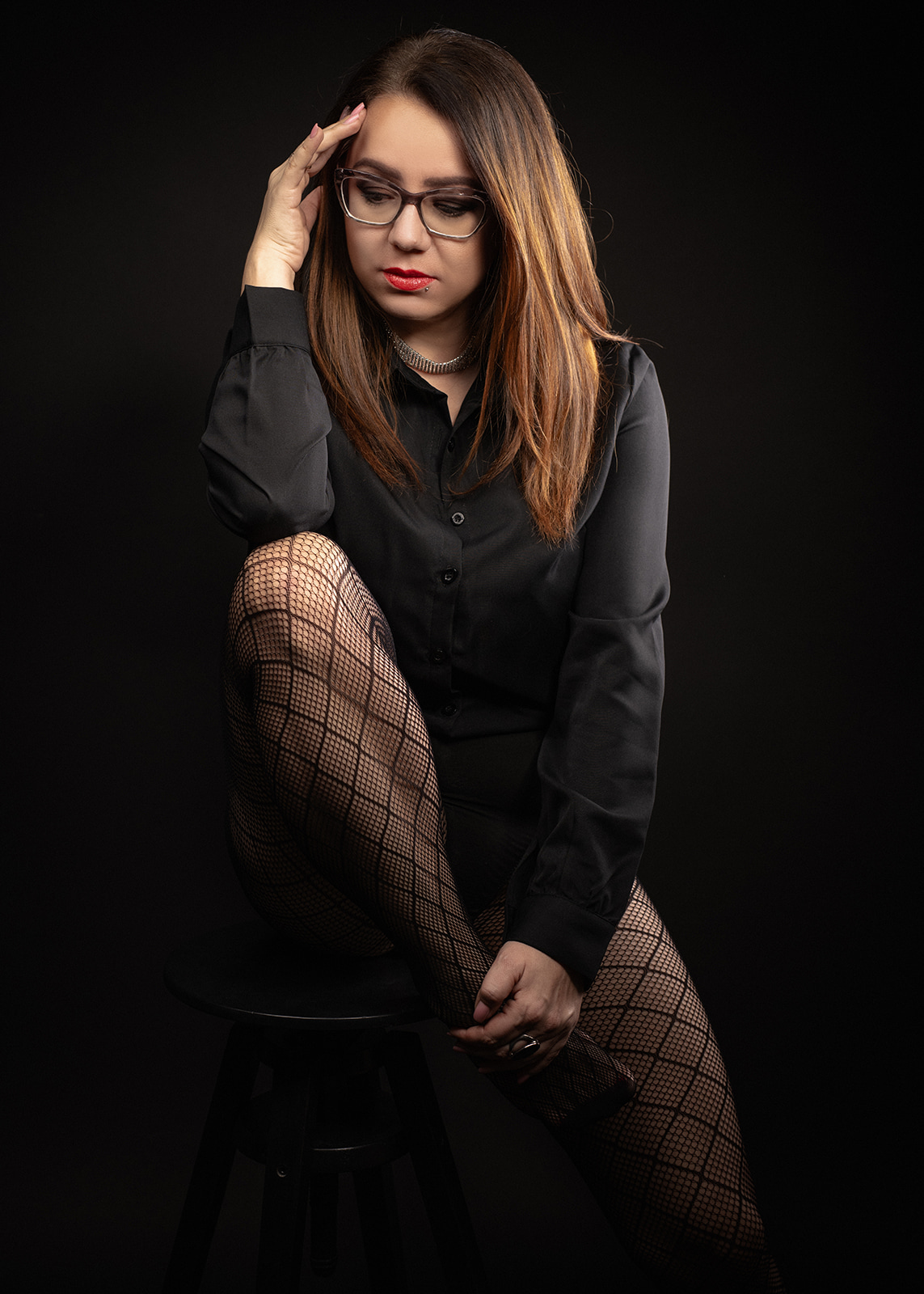 Woman wearing glasses and a black shirt seated against a dark studio background.