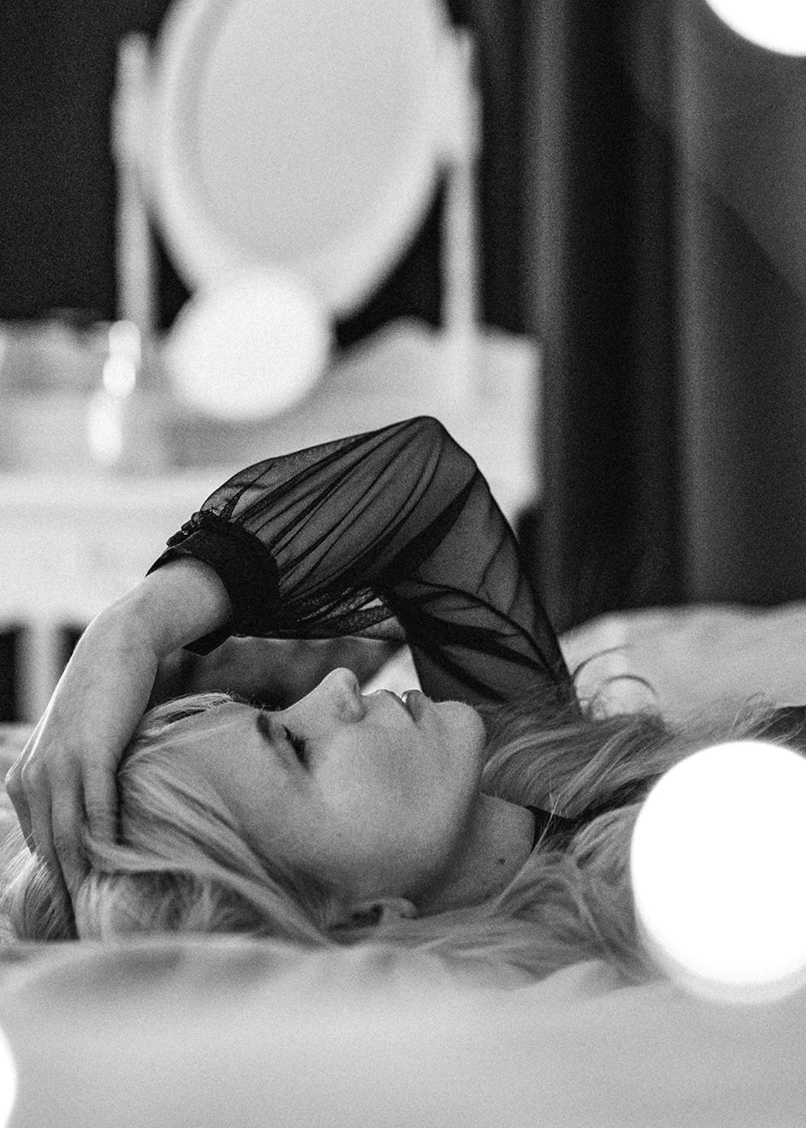 Black and white portrait of a blonde woman lying on a bed in soft light.